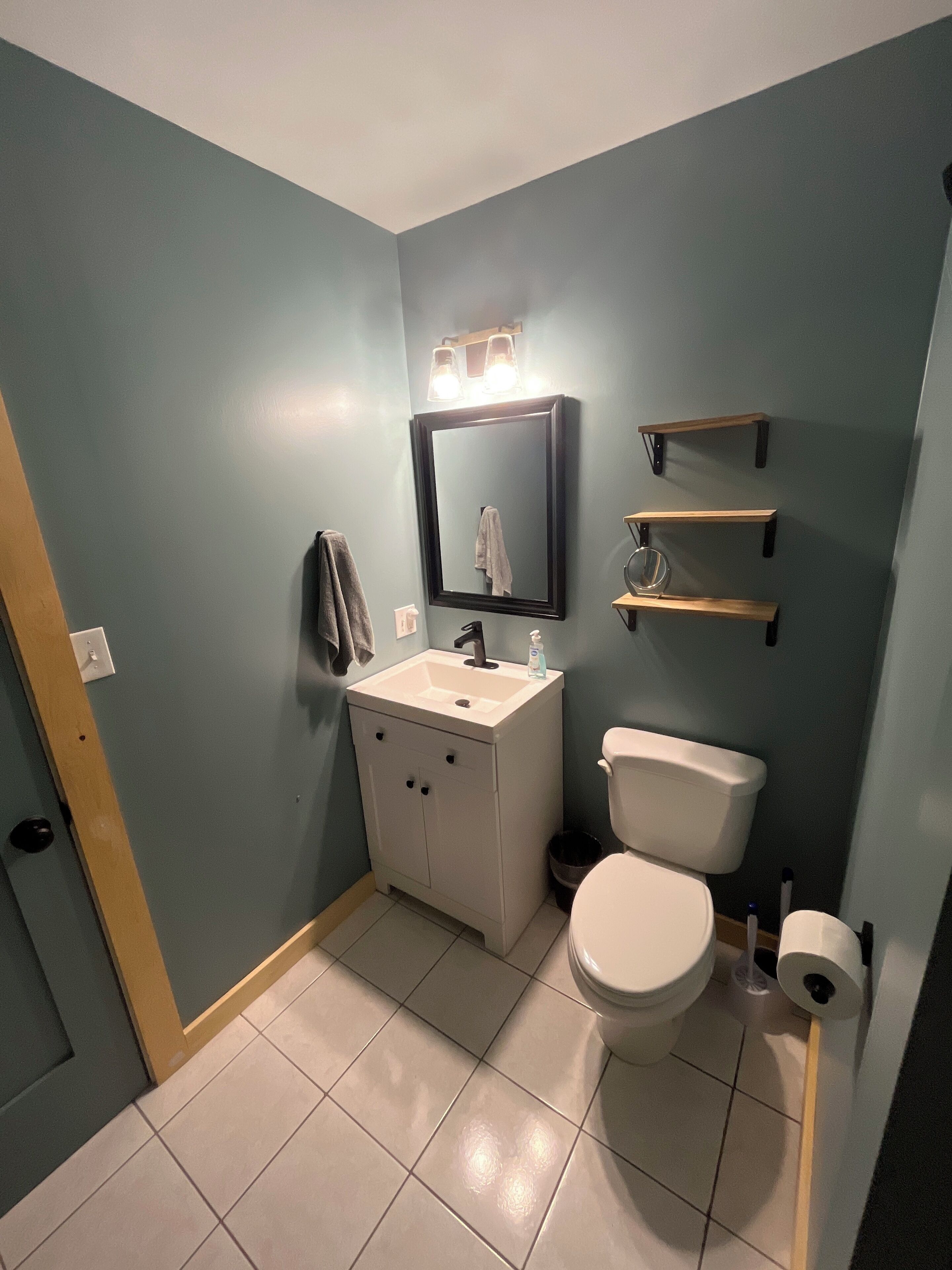 A bathroom with a white sink, a toilet, and a mirror. There are shelves above the toilet and a towel hanging on the wall.