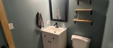 A bathroom with a white sink, a toilet, and a mirror. There are shelves above the toilet and a towel hanging on the wall.