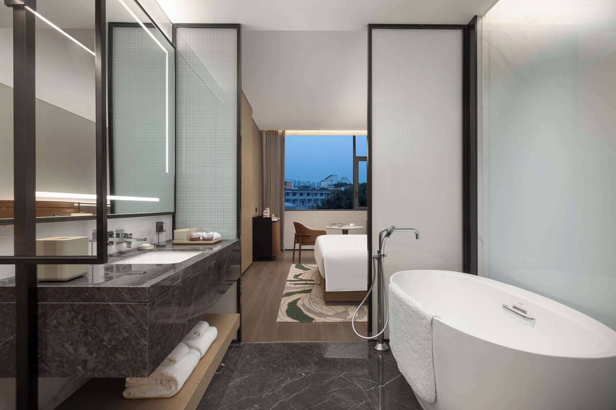 Ein modernes Badezimmer mit einem großen Spiegel, zwei Waschbecken, einer Badewanne und Blick auf ein Gebäude durch eine Glastür.