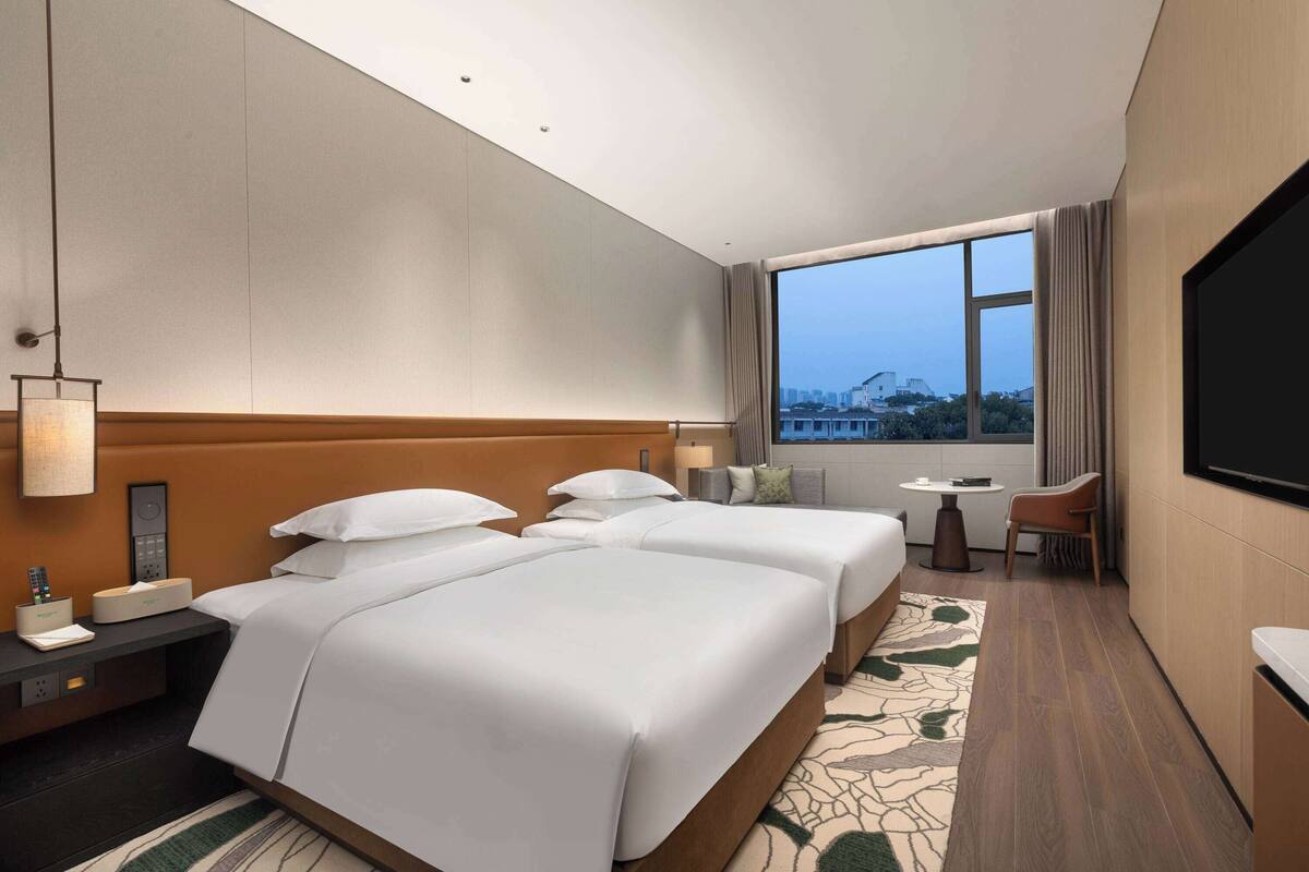 Ein Hotelzimmer mit zwei Betten, einem Fernseher, einem Schreibtisch und einem Fenster mit Blick auf die Stadt.