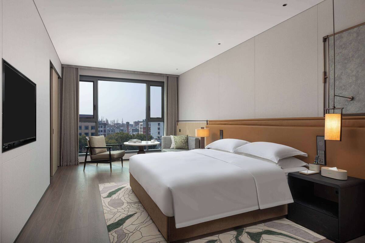 Ein modernes Hotelzimmer mit einem großen Bett, einem Flachbildfernseher, einem Schreibtisch mit Lampe und Blick auf die Stadt durch große Fenster.