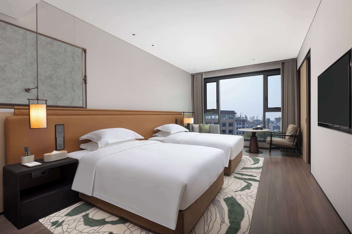 Ein Hotelzimmer mit zwei Betten, einem großen Fenster mit Blick auf die Stadt, einem Fernseher und einem Schreibtisch mit Lampe.