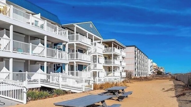 A beachfront hotel with multiple balconies, a sandy beach, and picnic tables.