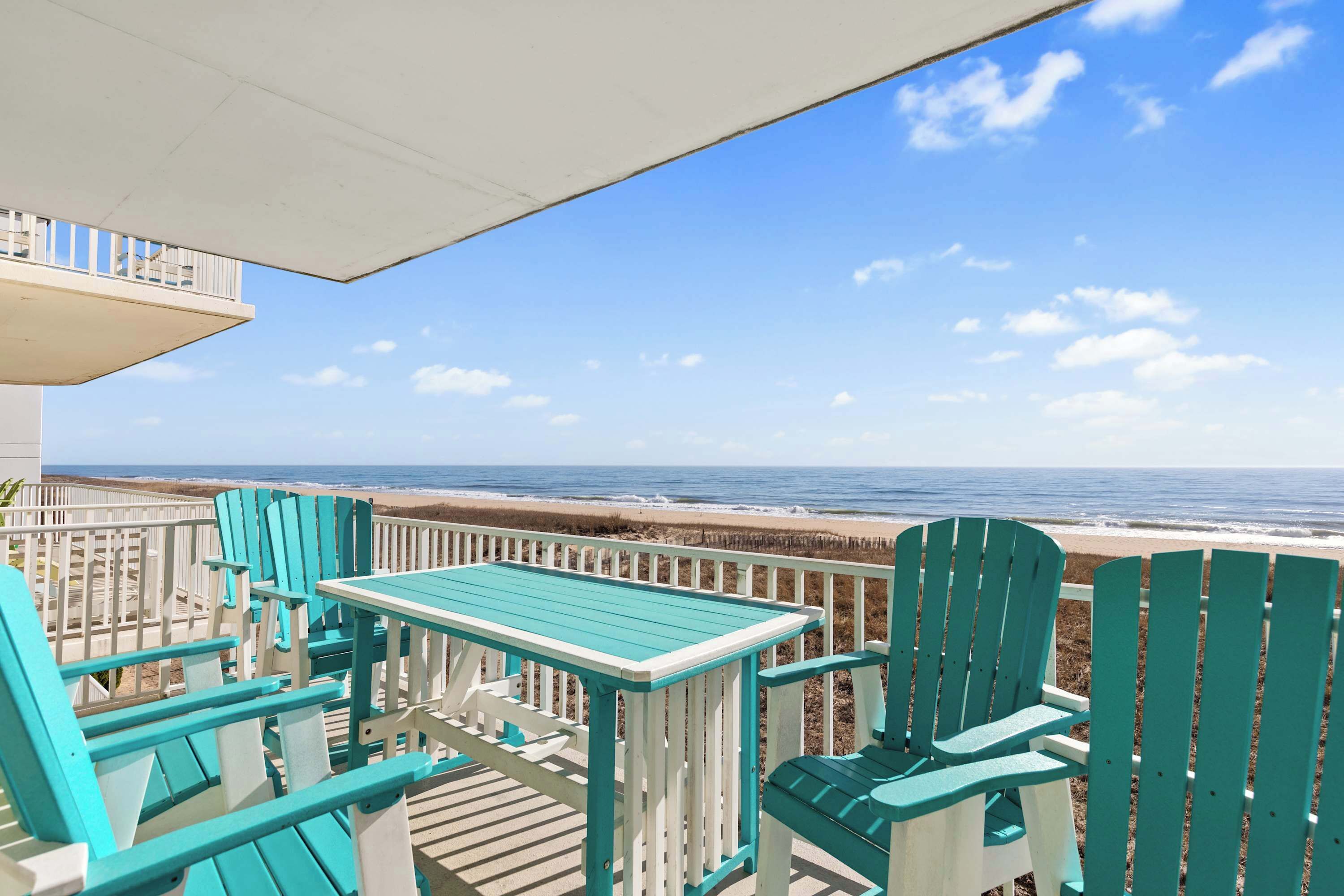 A deck with turquoise Adirondack chairs and a table overlooking a beach.