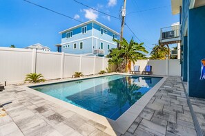 A rectangular swimming pool with a white fence surrounding it, a blue house in the background, and a clear blue sky.