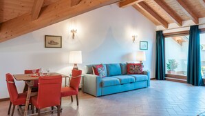 Ein gemütliches Wohnzimmer mit Holzbalkendecke, einem blauen Sofa, roten Sesseln, einem Esstisch mit Gläsern und einem Fenster mit Vorhängen.