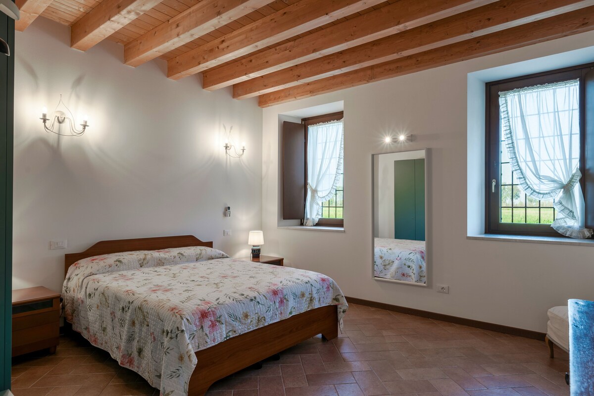 A bedroom with a wooden bed, two bedside tables, a nightstand, a window with sheer curtains, and a mirror above the window.