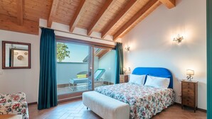 Ein Schlafzimmer mit Bett, einer Bank, einem Nachttisch, einer Wandlampe und einer Glastür zum Balkon.