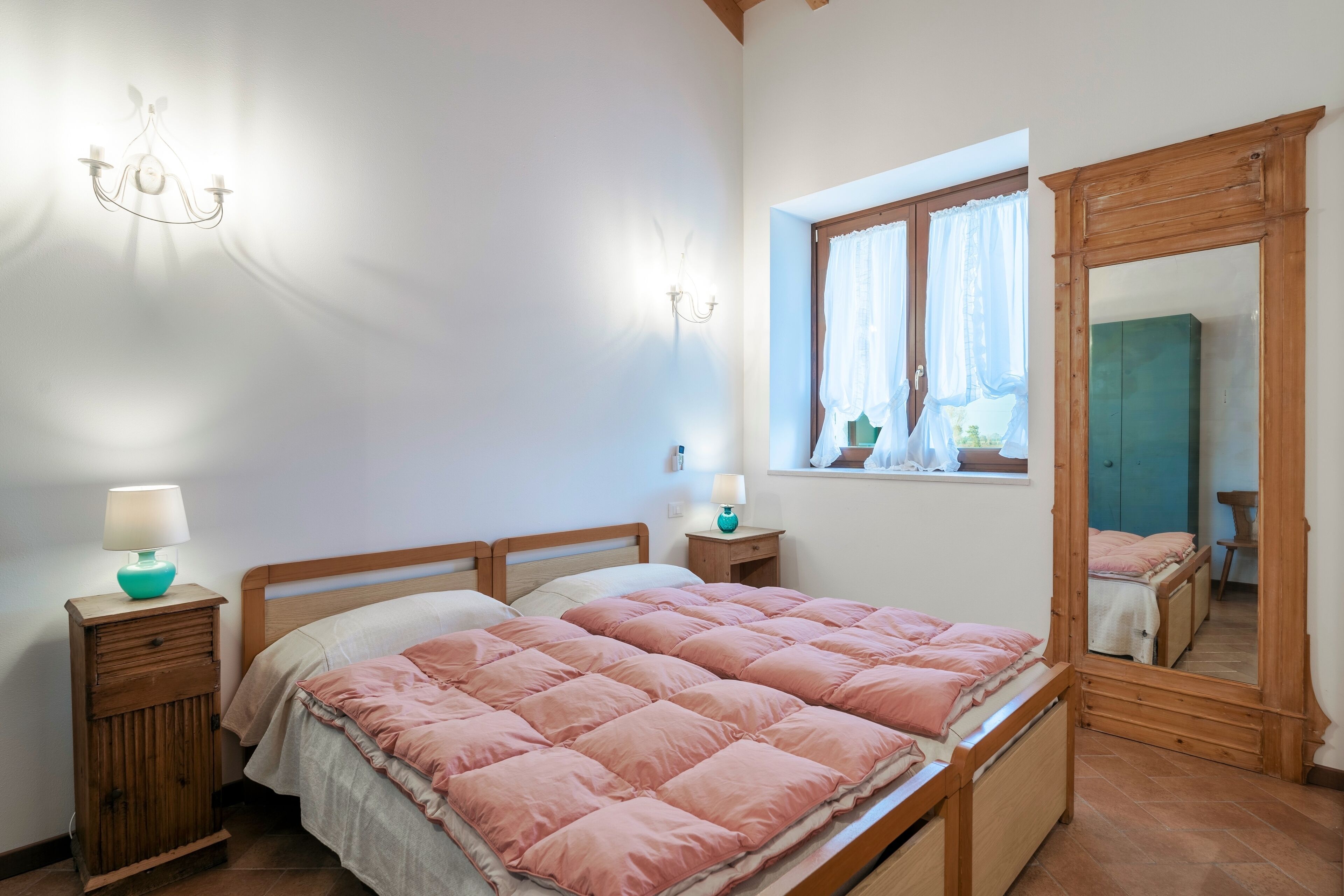 Un dormitorio con una cama de madera, una mesita de noche con lámpara, una ventana con cortinas y un espejo.