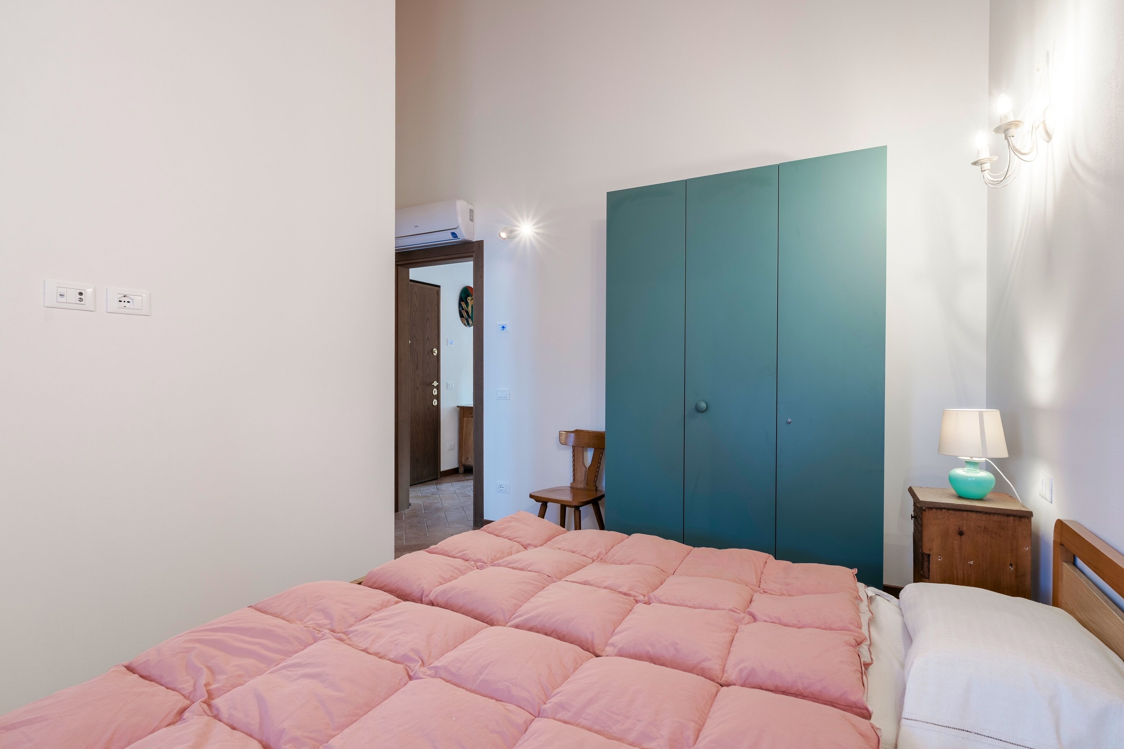A bedroom with a bed, a wardrobe, a chair, a nightstand with a lamp, and a wall socket.