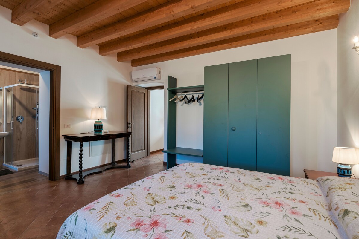 A bedroom with a bed, a wooden ceiling, a green wardrobe, a table with a lamp, and a bathroom with a shower.