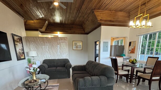 Un salón amplio con techo alto, vigas de madera y una lámpara moderna. Hay dos sofás grises, una mesa de centro de vidrio y un comedor con sillas de madera y una mesa de vidrio.