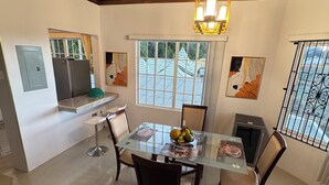 Comedor con mesa de vidrio, sillas y una lámpara de candelabro. Zona de cocina con refrigerador y ventana.