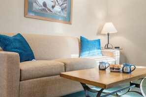 A room with a sofa, a small table with mugs, a lamp, and a framed picture on the wall.