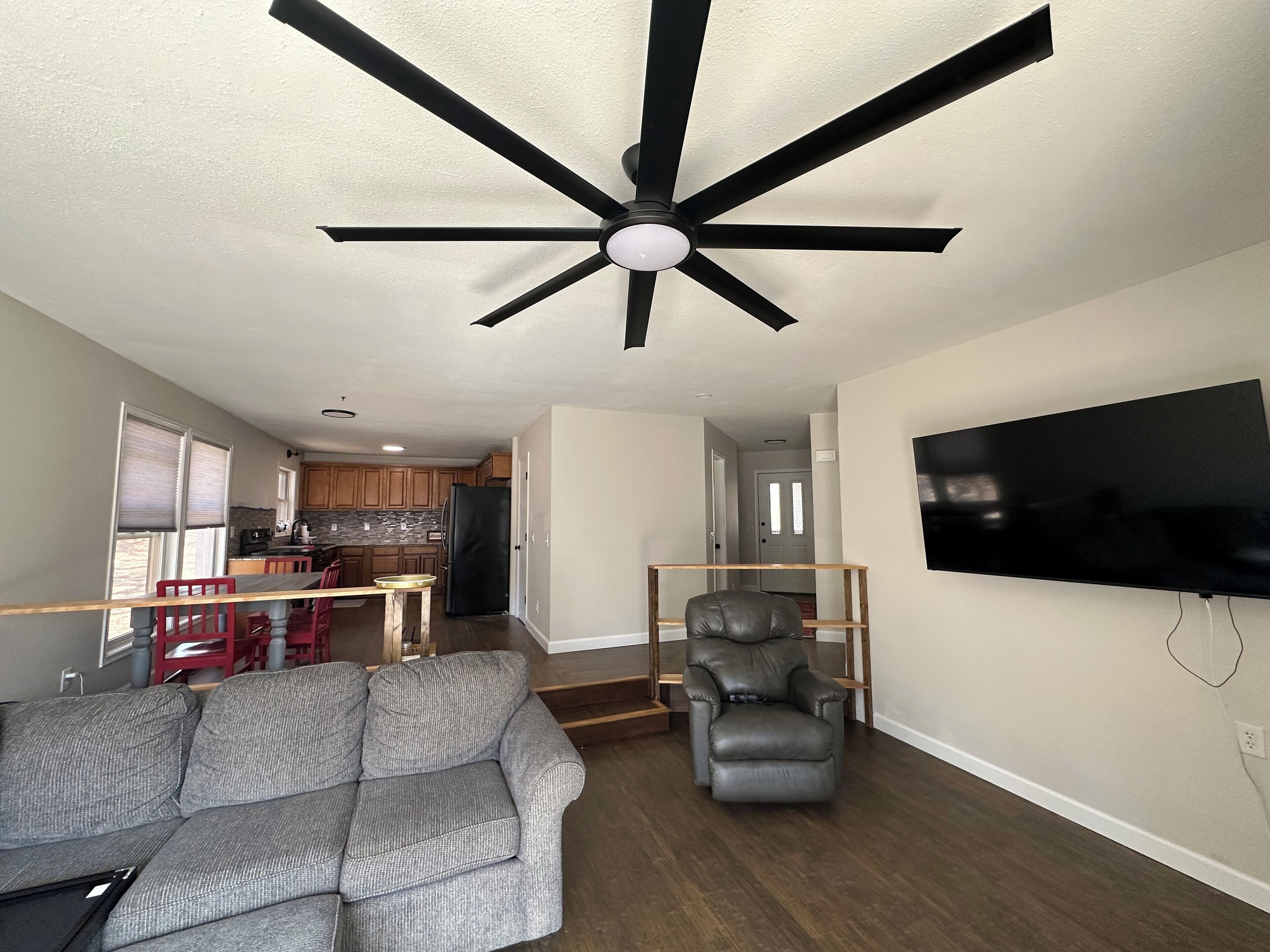 A living room with a gray sofa, a black leather chair, a ceiling fan, and a flat-screen TV mounted on the wall.