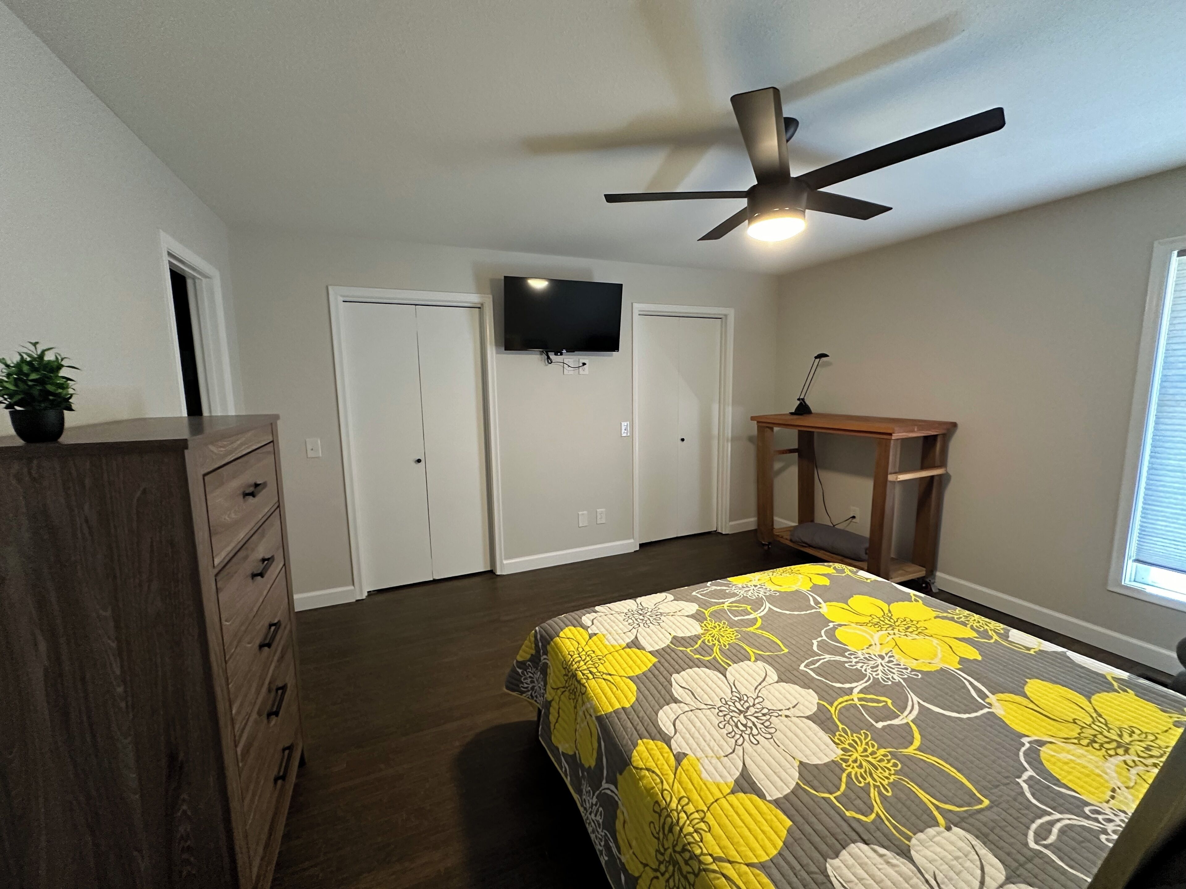 A bedroom with a bed, dresser, ceiling fan, TV, and a window.