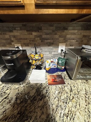Un comptoir de cuisine avec une machine à café Keurig, un support à dosettes de café, une pile d’essuie-tout, une boîte de stévia, une bouteille d’huile d’olive et une multiprise Black & Decker.