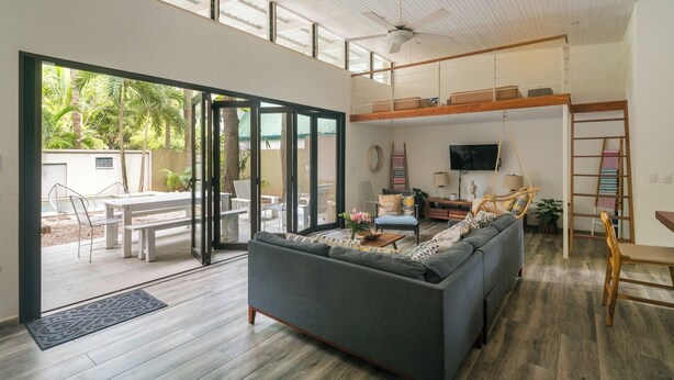 A modern living room with a grey sofa, a wooden coffee table, and a TV on the wall. There is a balcony with a wooden railing and a view of greenery outside. - Oceano5, 4 bedroom Pool Villa - 4 min walk to the beach (nosara)