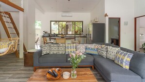 A modern living room with a grey sectional sofa, a wooden coffee table, and a wicker chair. A kitchen is visible in the background with a large window. - Oceano5, 4 bedroom Pool Villa - 4 min walk to the beach (nosara)