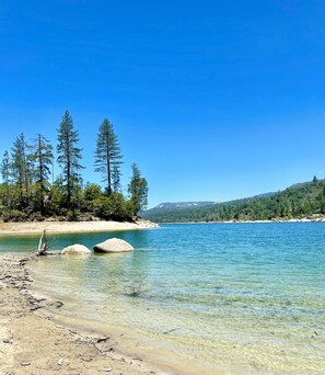A clear lake with a sandy shore, surrounded by trees and hills in the background.