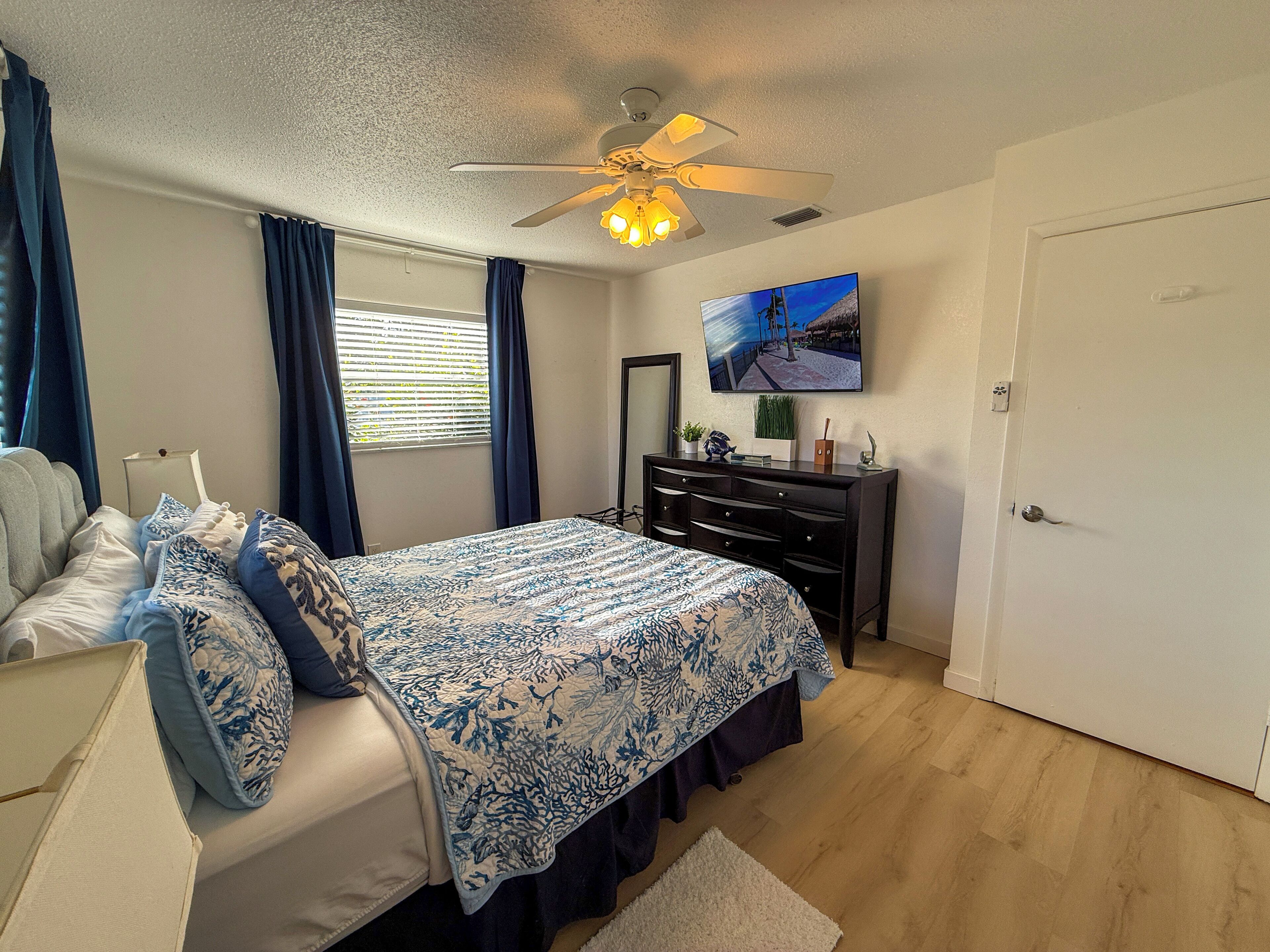 Une chambre à coucher comprenant un lit, une commode, un ventilateur de plafond, une fenêtre avec des rideaux, un téléviseur fixé au mur et une porte.