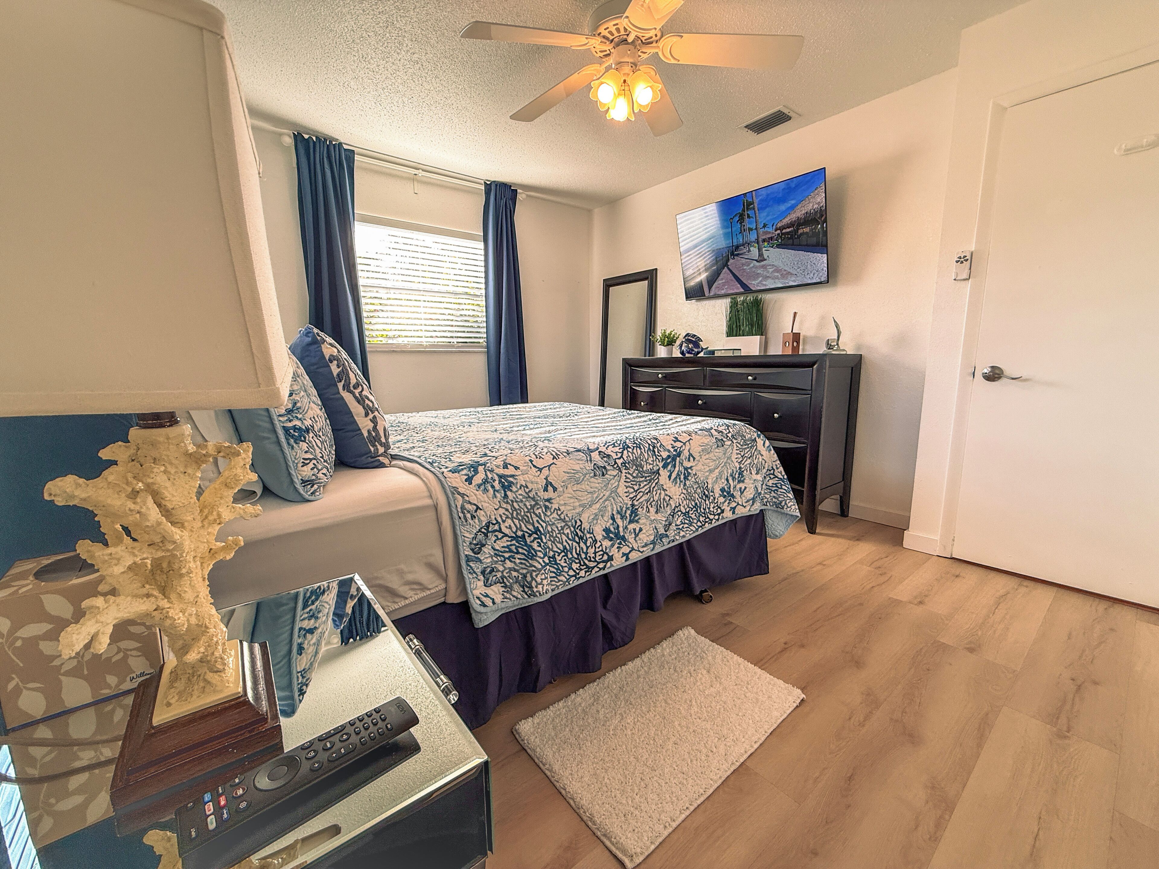 A bedroom with a bed, a dresser, a television on the wall, and a ceiling fan.