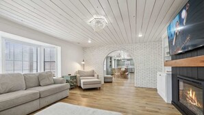 Een moderne woonkamer met een open haard, een bank, een eethoek en een keuken, zichtbaar door een boog.