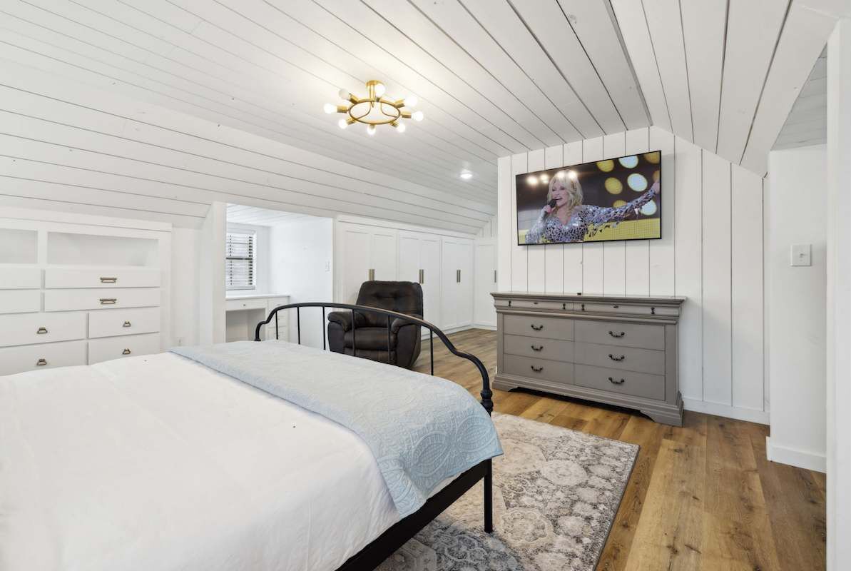 Um quarto com cama, cômoda, cadeira, televisão na parede e luminária no teto.