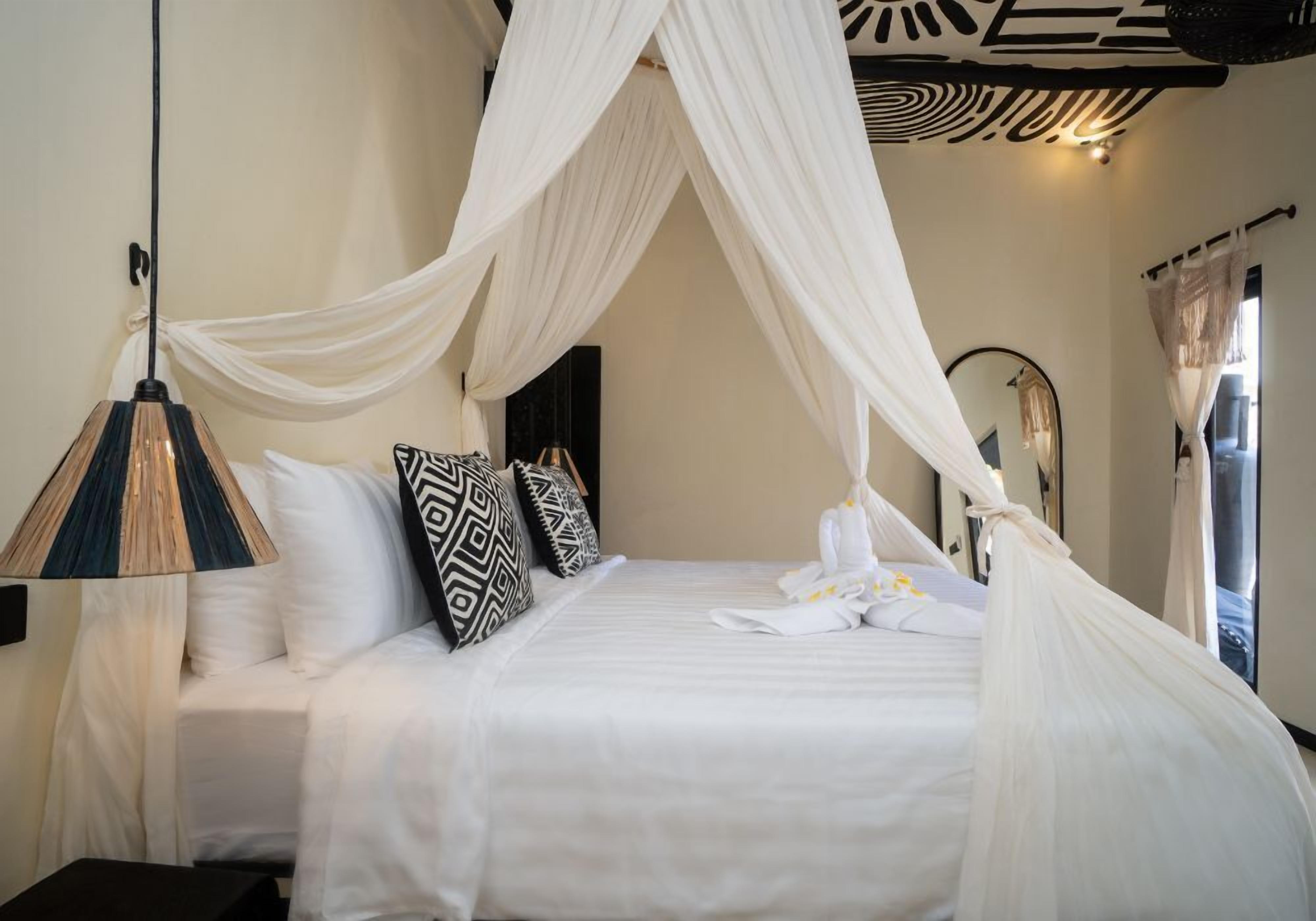 A neatly made bed with white bedding and a canopy, a bedside lamp, and patterned pillows.