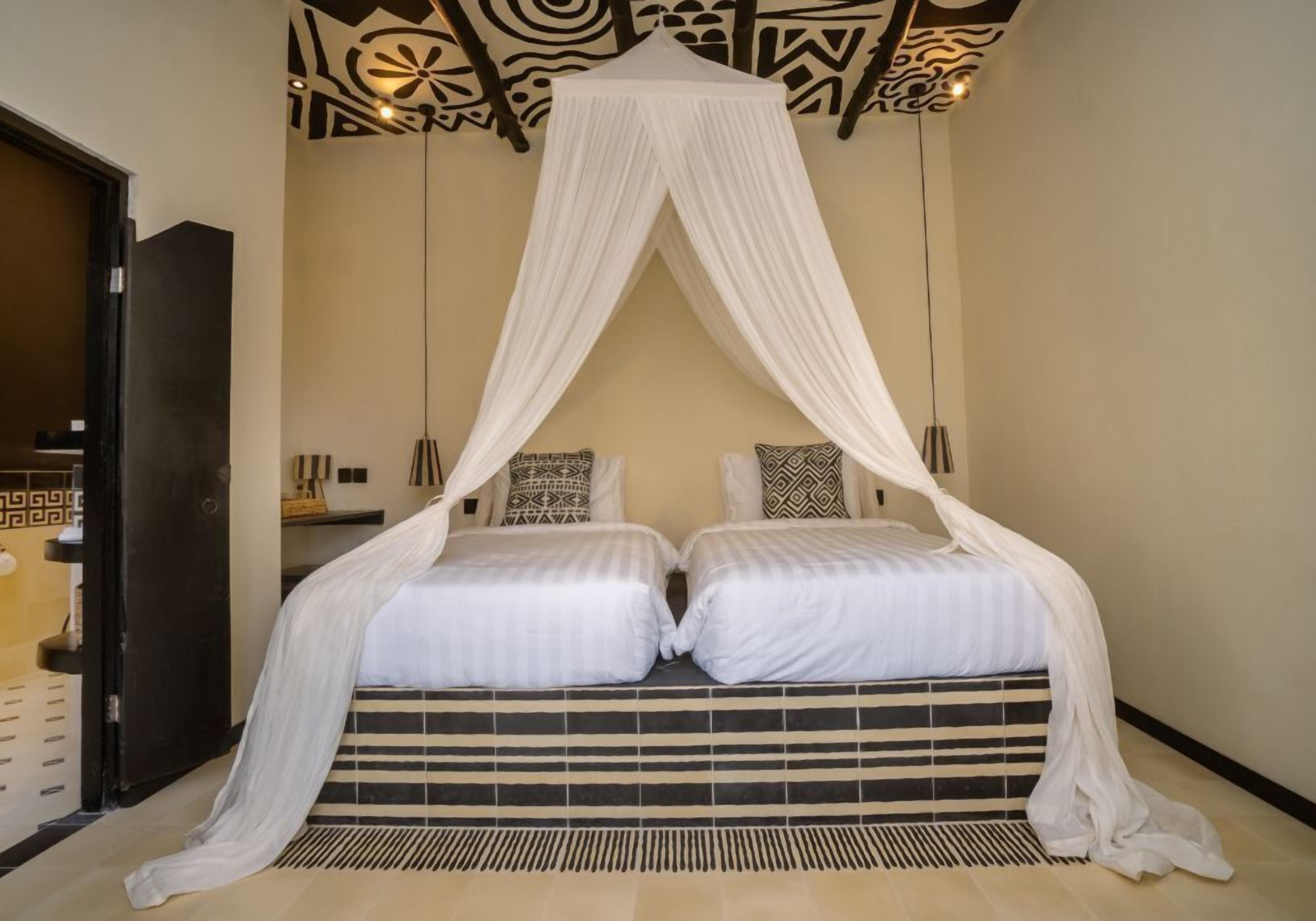 A room with a bed under a white canopy, two pillows with geometric patterns, a black and white striped bed runner, and a patterned ceiling.