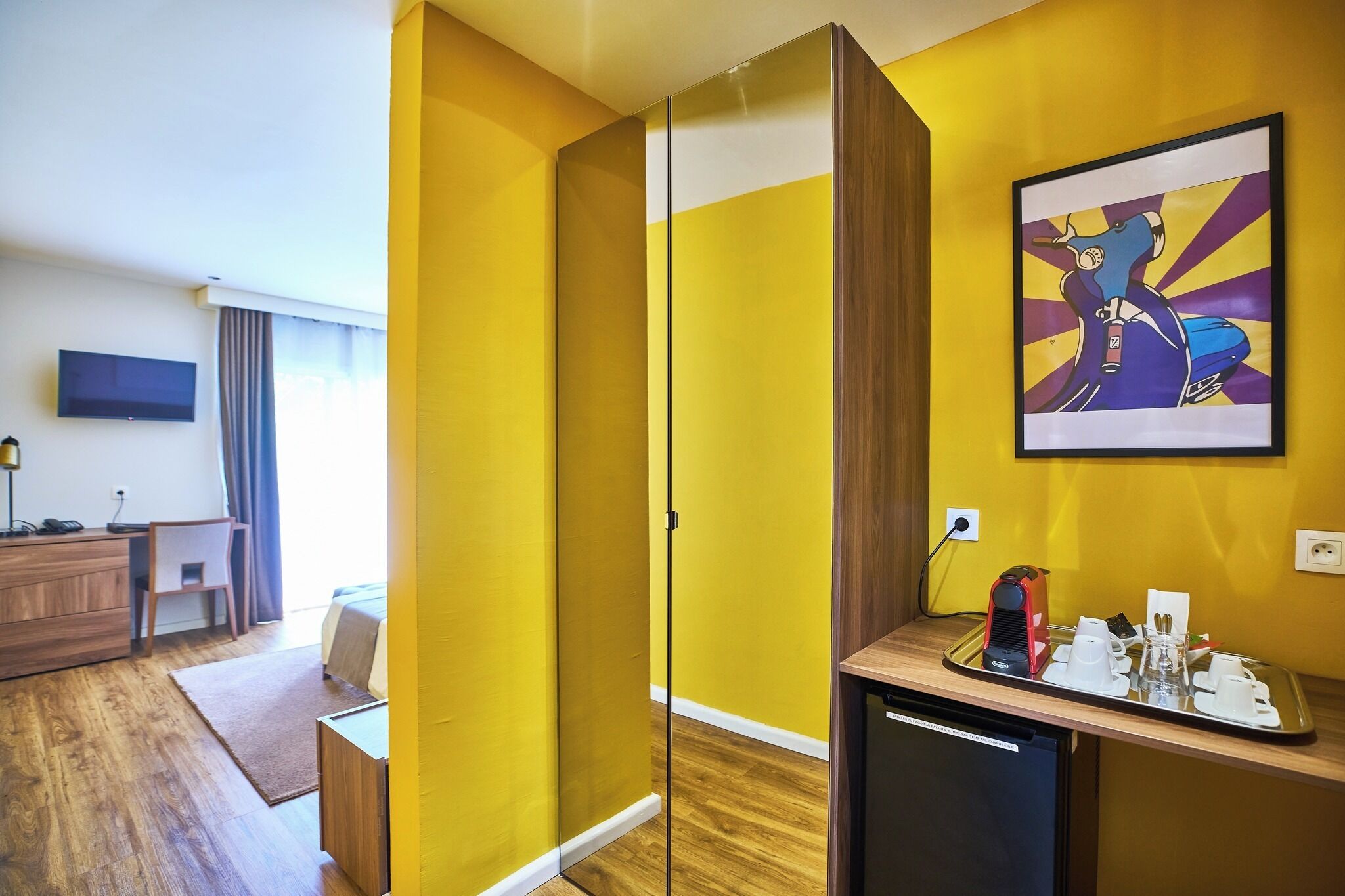 Quarto de hotel com parede amarela, uma cama, escrivaninha com cadeira, televisão e um quadro.