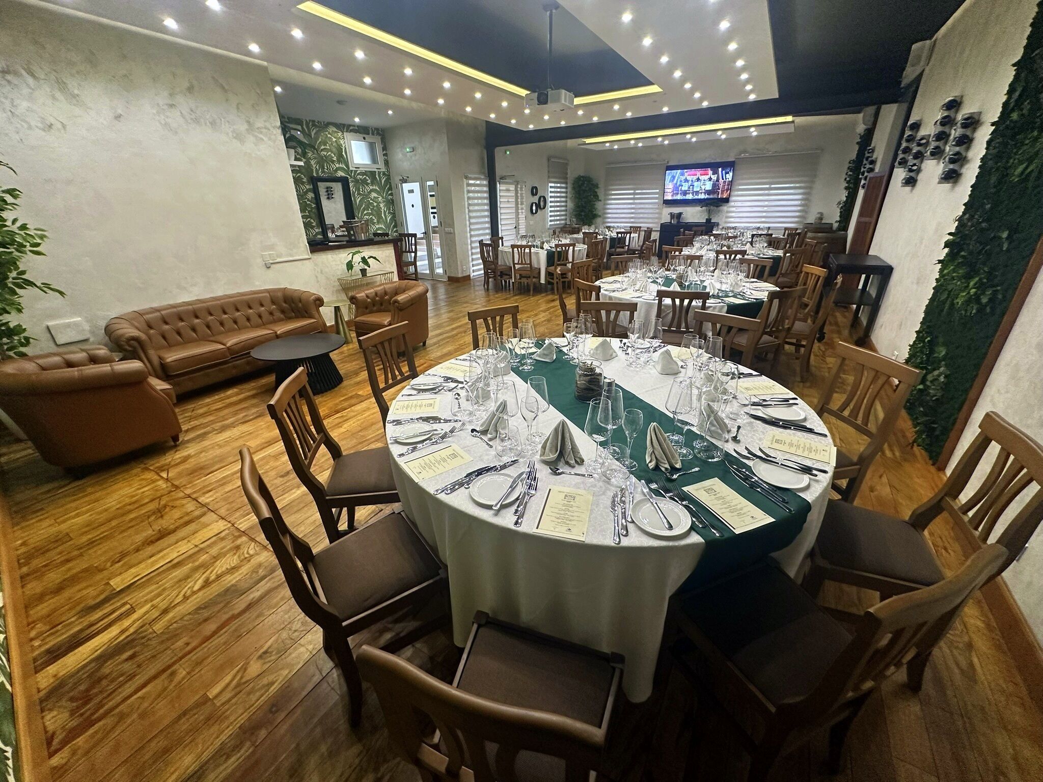 A restaurant with round tables set for dining, wooden flooring, and a green wall with plants.
