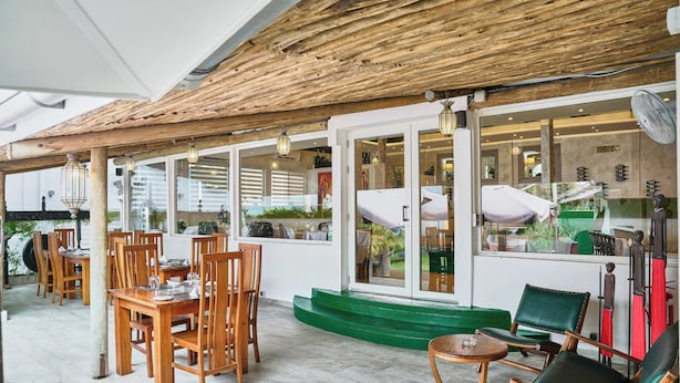 An outdoor patio with wooden tables and chairs, a green cushioned seating area, and a fan. - Roma Hôtel & Restaurant (Libreville)