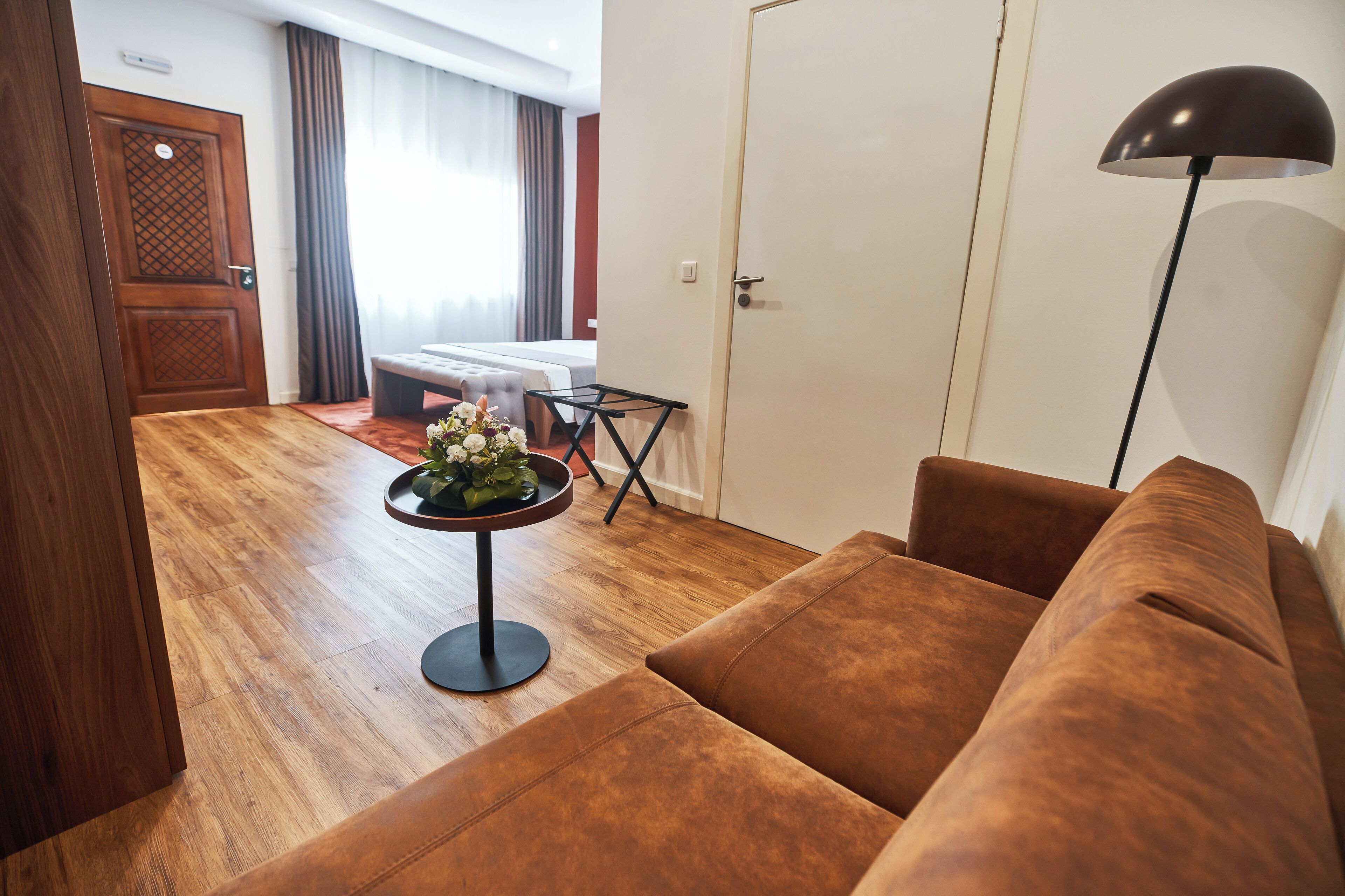 A hotel room with a wooden floor, a brown sofa, a small round table with a flower arrangement, a bed with a white headboard, and a door with a decorative panel.