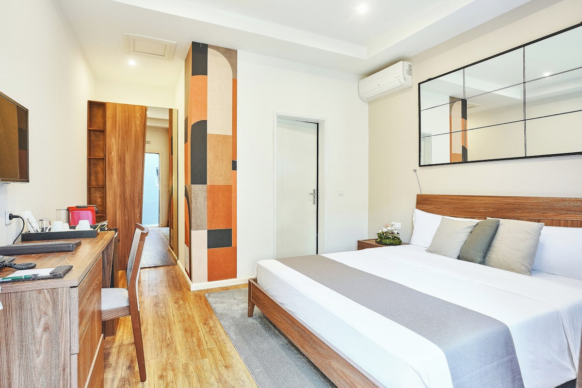 Ein modernes Hotelzimmer mit Schreibtisch aus Holz, einem Bett mit grauer Bettdecke, einem großen Spiegel und einem Kleiderschrank.