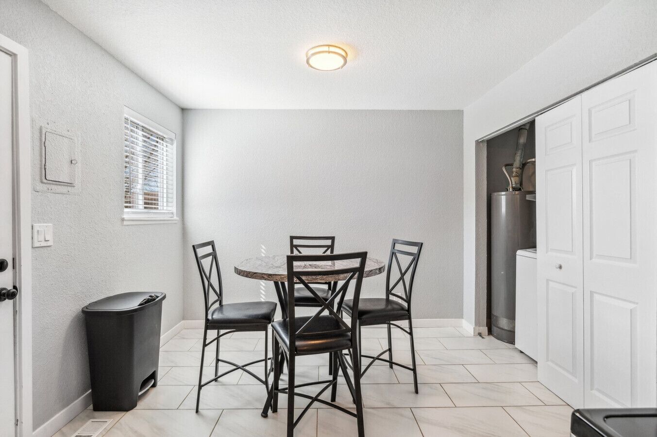 A compact kitchen with a round dining table and four chairs, a trash bin, a window with blinds, and a white door.