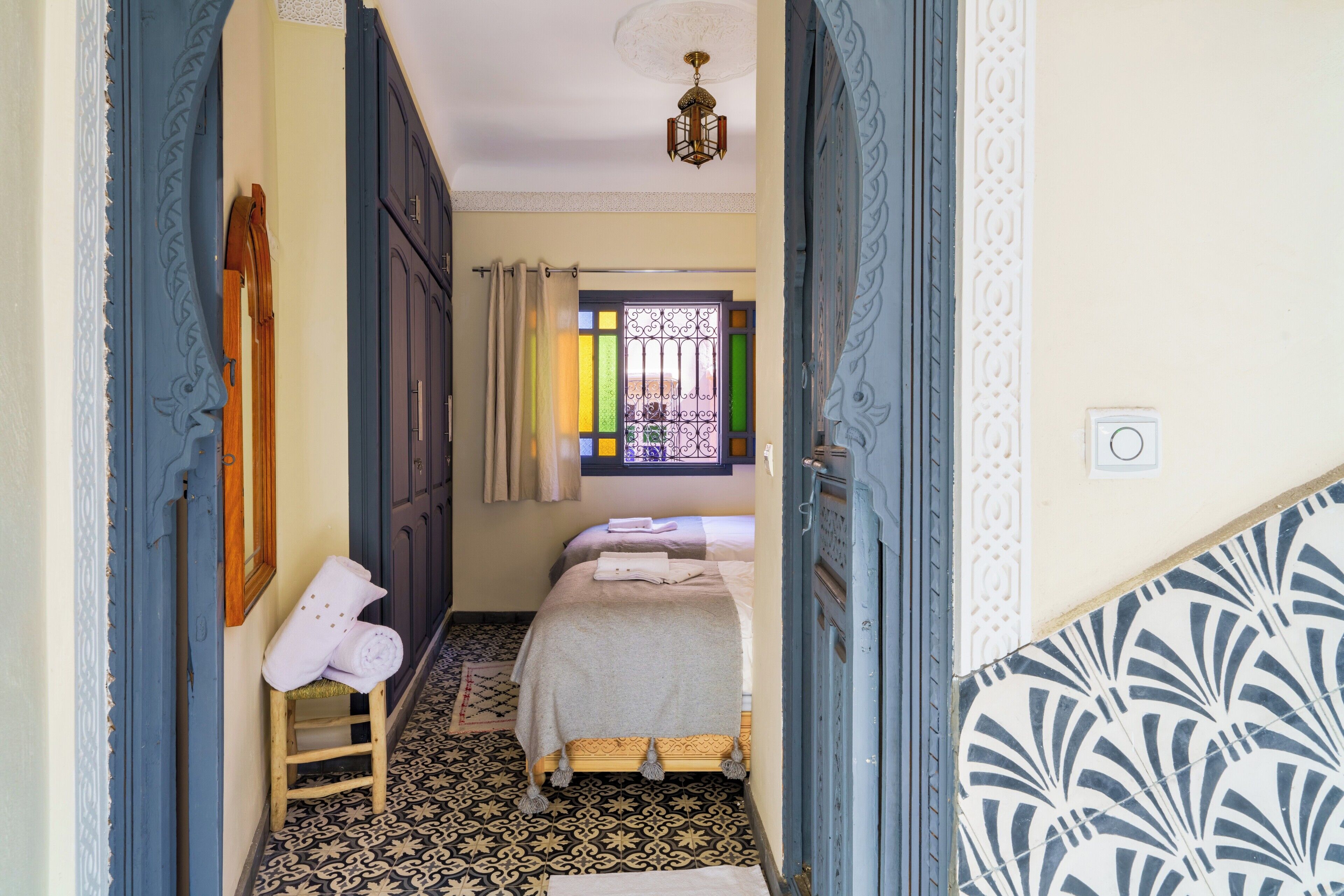 Un couloir étroit mène à une chambre où se trouvent un lit, un tabouret en bois et une fenêtre ornée de barreaux décoratifs.
