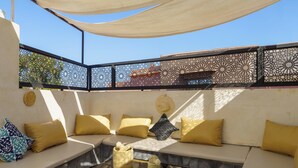 Un espacio al aire libre, iluminado por el sol, con una zona de estar que incluye un sofá y una mesa de centro de vidrio, una cerca decorativa y una cesta tejida.