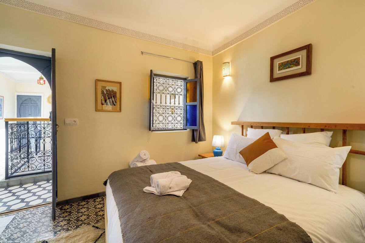 A hotel room with a bed, a nightstand, a window with a curtain, a door, and a view of the outside balcony.