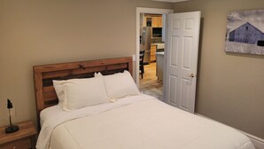 Un dormitorio con cabecera de madera, cubrecama blanco y una puerta que da a la cocina.