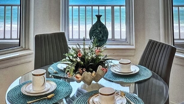 Un coin repas avec une table dressée pour quatre personnes, un vase de fleurs et une vue sur l’océan grâce à de grandes fenêtres.