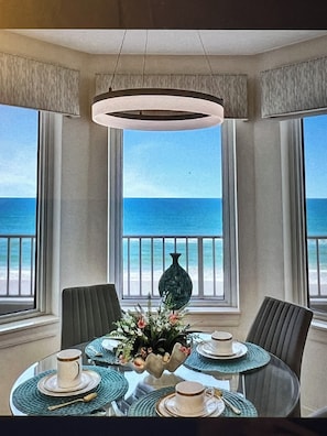 A dining area with a table set for four, a vase of flowers, and a view of the ocean through large windows.