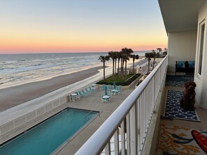 A balcony with a view of the beach, a pool, and a dog.