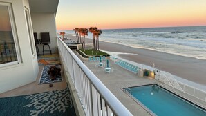 Un balcon avec vue sur la plage et l’océan, une piscine et un chien allongé sur un tapis.
