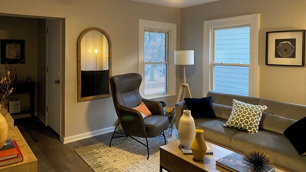 Un salon avec un ventilateur de plafond, un canapé avec des coussins, une table d’appoint avec un vase et des livres, et un miroir avec une lampe.
