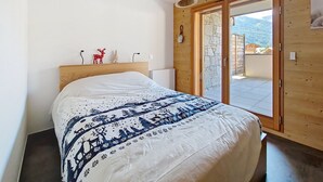 Une chambre avec une tête de lit en bois, un lit recouvert d’une couette à motifs, une petite décoration représentant un renne rouge, une table de chevet en bois et une vue sur un paysage montagneux par une porte vitrée.