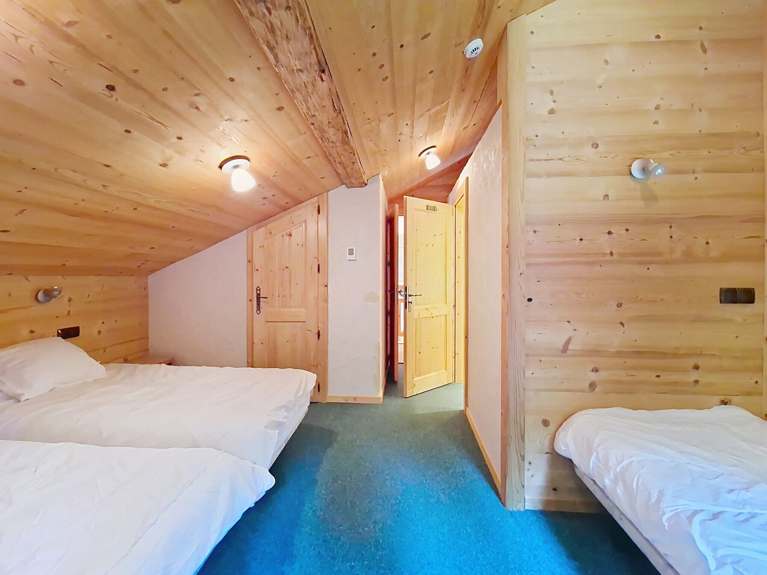 Een kamer met houten wanden en plafond, twee bedden met wit beddengoed, een blauw vloerkleed en een deur naar een andere kamer.