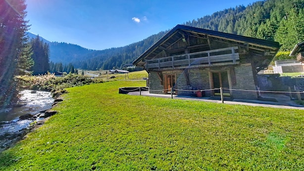 Une cabane en bois, construite sur des fondations en pierre, entourée d’une zone herbeuse et bordée d’un ruisseau.