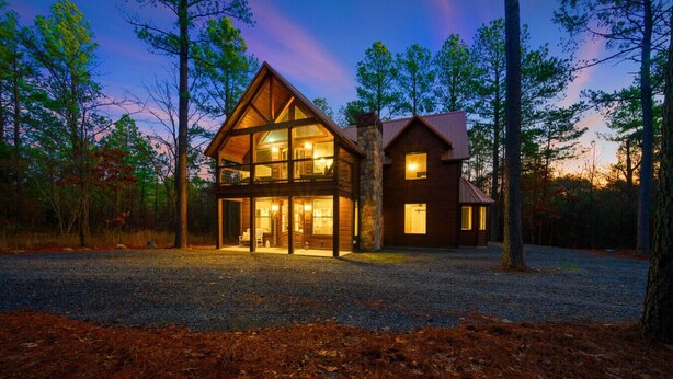 A two-story log cabin with a stone foundation and large windows, surrounded by a forested area.