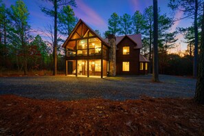 A two-story log cabin with a stone foundation and large windows, surrounded by a forested area.